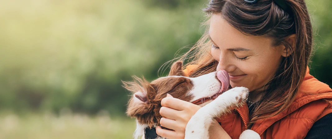 Mobile_About_Us_What_Were_Building A woman in an orange vest smiles as a brown and white dog licks her face. She hugs the dog lovingly outdoors, with a blurred green background.
